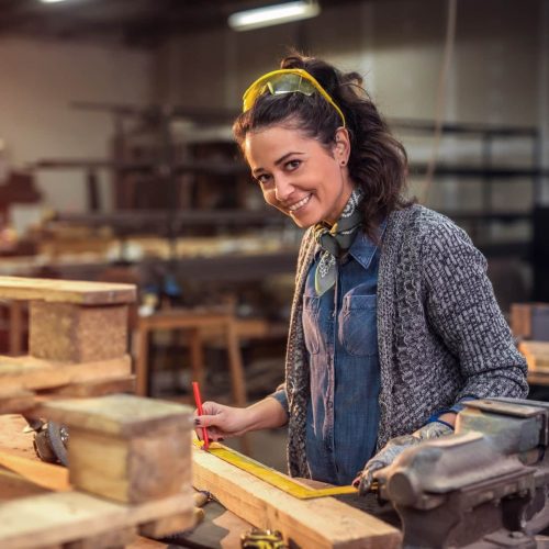 Middle aged professional female carpenter looking at the camera while taking measures of raw wood in her workshop.